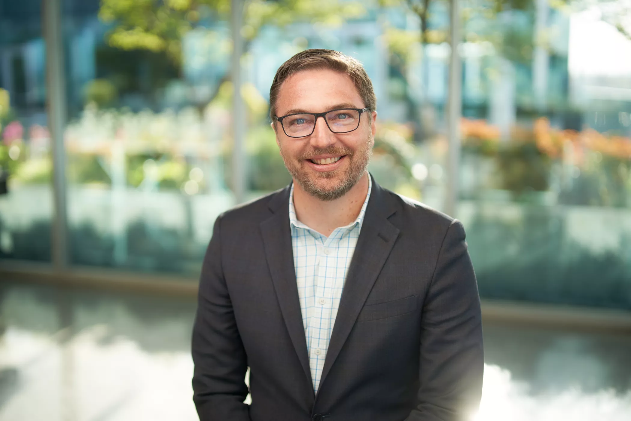 A person wearing a dark suit and light checkered shirt is seated in a bright, modern office environment. The background features large glass windows with greenery and soft natural light. The setting conveys a professional and contemporary atmosphere.